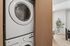 Modern apartment laundry area with sleek stacked washer and dryer near a bright living space, accented by a stylish potted plant.