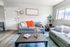Bright and welcoming living room with modern gray sofas, vibrant orange accents, and lush greenery by a sunlit window.
