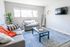 Bright and inviting living room featuring cozy gray sofas, vibrant orange throw blanket, stylish coffee table, and large window with natural light.