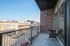 Modern urban apartment balcony overlooking lively street, featuring cozy outdoor seating and glass doors for natural light.