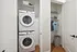 Modern laundry area with stacked washer and dryer, adjacent to a neatly organized closet space with hanging clothes and shelves, complemented by elegant wood flooring for a sleek apartment look.