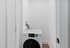 Modern laundry space featuring a sleek, front-loading washer in a compact setting with white walls and hardwood flooring.