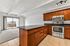 Modern apartment kitchen with stainless steel appliances, sleek wooden cabinetry, and a spacious granite countertop overlooking a bright living area with floor-to-ceiling sliding glass doors and city views.