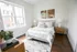 Bright and serene bedroom featuring a plush bed with soft linens, large windows allowing natural light, modern decor, and wooden furniture.