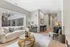 Bright and modern living space featuring a cozy sofa, stylish wicker coffee table, and a compact home office corner with a desk and chair, looking out to large windows with a pleasant urban view.