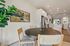 Bright dining area with elegant round table, modern chairs, lush plant, and scenic artwork overlooking a stylish white kitchen with stainless steel appliances.