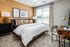 A comfortable bedroom featuring a cozy bed with plush bedding, a nightstand with a lamp, and a large window allowing natural light to fill the room at Abberly Alston Apartments in Cary, NC
