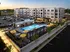 Modern apartment complex featuring a luxurious swimming pool with vibrant yellow umbrellas, surrounded by lush landscaping and ample parking.