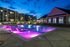 Luxurious apartment complex at dusk featuring an illuminated pool with vibrant purple lighting, surrounded by comfortable lounge chairs and modern architecture.
