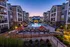 Luxurious apartment complex featuring a sparkling central pool, cozy patio spaces with umbrellas, and modern multi-story buildings at sunset.
