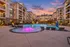 Captivating evening view of a modern apartment complex with a sparkling pool and glowing purple fountain, set against a picturesque sunset sky.