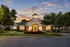 Elegant apartment building with a warmly lit entrance, surrounded by lush gardens and trees, set against a stunning sunset sky.