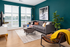 Chic living room with a modern gray sofa, vibrant teal walls, and ample natural light from large windows.