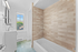 Bright and modern bathroom featuring elegant beige tiles, a sleek white bathtub, and a large window offering natural light.