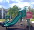 Bright playground with slides and climbing structures beside residential buildings under a blue sky.