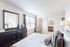 Bright and spacious bedroom with elegant decor, featuring a large mirror, stylish dresser, and cozy seating next to a window, leading to an ensuite bathroom.