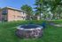 Charming brick apartment complex with lush green lawns, featuring a cozy outdoor fire pit and inviting blue Adirondack chairs set amongst mature trees.