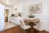 Charming dining area with a round wooden table, elegant chairs, and a plant centerpiece, adjacent to a cozy kitchen with white cabinetry.