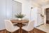 Charming dining area with a round wooden table, modern chairs, and a potted plant, adjacent to a bright kitchen with white appliances.