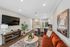Inviting open-concept living space with modern decor, featuring a plush rust-colored sofa, a stylish round coffee table, and a sleek wall-mounted TV. Cozy kitchen area with high-back woven chairs and trendy black backsplash.