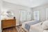 Charming bedroom with elegant neutral decor, featuring a cozy bed, a stylish wooden dresser with a chic lamp, and soft natural light from large windows.
