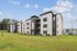 Modern apartment complex with contemporary architecture set against a bright blue sky, surrounded by lush green landscaping.