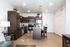 A modern open-concept kitchen and entryway featuring dark wood cabinetry, speckled granite countertops, stainless steel appliances, and wood-style flooring. A kitchen island sits in the center with two dark wooden bar stools tucked under it, illuminated by pendant lights above. To the right is a white front door alongside white double doors, and in the foreground on the left, a portion of a living room is visible with a lamp and side table.