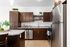 A modern open-concept kitchen and entryway featuring dark wood cabinetry, speckled granite countertops, stainless steel appliances, and wood-style flooring. A kitchen island sits in the center with two dark wooden bar stools tucked under it, illuminated by pendant lights above. To the right is a white front door alongside white double doors, and in the foreground on the left, a portion of a living room is visible with a lamp and side table.
