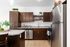 A modern open-concept kitchen and entryway featuring dark wood cabinetry, speckled granite countertops, stainless steel appliances, and wood-style flooring. A kitchen island sits in the center with two dark wooden bar stools tucked under it, illuminated by pendant lights above. To the right is a white front door alongside white double doors, and in the foreground on the left, a portion of a living room is visible with a lamp and side table.