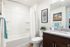 A modern apartment bathroom featuring a dark wood vanity with a speckled granite countertop, undermount sink, and large wall mirror. Adjacent sits a white toilet beneath framed wall art and a small decorative plant, beside a white tub-and-shower combo with a curved curtain rod, white shower curtain, and teal and white towels hanging nearby.