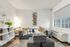 Cozy living room interior at Fusion at Rye Apartments in Houston Texas featuring a gray sofa with fluffy pillows, stacked ottoman cubes, wall hanging shelves with small plants, a white shelving unit with decor pieces, a side table with a modern lamp, large window with a roller shade, in room air conditioning unit, and wood style flooring.