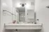 Bathroom vanity at Fusion at Rye Apartments in Houston Texas showing a wide marble style countertop, chrome faucet, soap dispensers, a mounted medicine cabinet, wall mounted towel bars, an overhead light fixture with two bulbs, and a view of the shower area reflected in the mirror.
