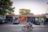 Charming row of local shops with colorful facades, a cyclist passing by, and cars parked in front, set against a backdrop of leafy green trees.