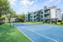 Charming apartment complex with modern balconies adjacent to a lush green tennis court under a vibrant blue sky.