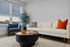 Chic living room with modern white sofa, vibrant orange cushion, elegant blue armchair, and lush green plant, bathed in natural light from large windows.