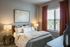 Inviting bedroom with modern decor, featuring a plush bed adorned with stylish cushions, a vibrant abstract painting, and warm natural lighting through elegant drapes.