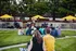 Vibrant outdoor concert in a lush park setting with yellow umbrellas, colorful flowers, and a lively audience.