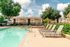 Inviting apartment community pool area with lounge chairs, surrounded by lush greenery and charming residential buildings under a bright blue sky.