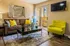 Inviting living room with a plush brown sofa adorned with vibrant yellow pillows, a sleek coffee table, and a cheerful yellow armchair, complemented by stylish decor and a wall-mounted TV next to a bright open patio door.