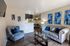 Elegant living room with plush grey sofas, blue accent chairs, and abstract blue wall art, featuring a modern glass coffee table and hardwood floors, leading to a cozy dining area.