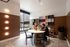 Modern apartment interior with a cozy seating area, featuring two people at a round table, large windows letting in natural light, and an artistic wall display. The space is accented with stylish lighting and a contemporary rug.