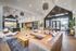 Modern living space with high ceilings, featuring a stylish leather sectional, rustic coffee table, and chic lighting. Open concept design with expansive windows offering natural light and a view of the outdoors.