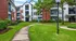 Charming apartment complex with brick and white facades, surrounded by lush green lawns and tall trees, perfect for a serene lifestyle.