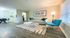 Modern apartment interior featuring an elegant living room with a stylish gray sofa, chic blue accent chair, and a cozy area rug. Bright natural light illuminates the open-concept space, leading to a dining area in the background.