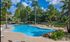 Luxurious apartment complex featuring a large, sparkling swimming pool surrounded by lush palm trees and relaxing lounge chairs, under a vibrant blue sky.