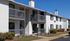 Charming two-story apartment building with white brick exterior, private balconies, and lush landscaping under a clear blue sky.