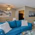 Chic living room with vibrant blue sofa, elegant artwork, and modern ceiling fan, adjacent to a cozy kitchen.