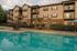 Charming apartment complex featuring three-story buildings with private balconies, surrounded by lush greenery and a sparkling swimming pool with relaxing lounge chairs and umbrellas.