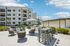 Modern apartment complex with stylish outdoor seating area, featuring comfortable chairs and a sleek barbecue setup, perfect for social gatherings under a clear blue sky.