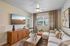 Bright and inviting living room featuring a chic white sectional sofa, modern wooden coffee table, stylish entertainment console, and large TV. Abundant natural light with lush outdoor views through wide windows.